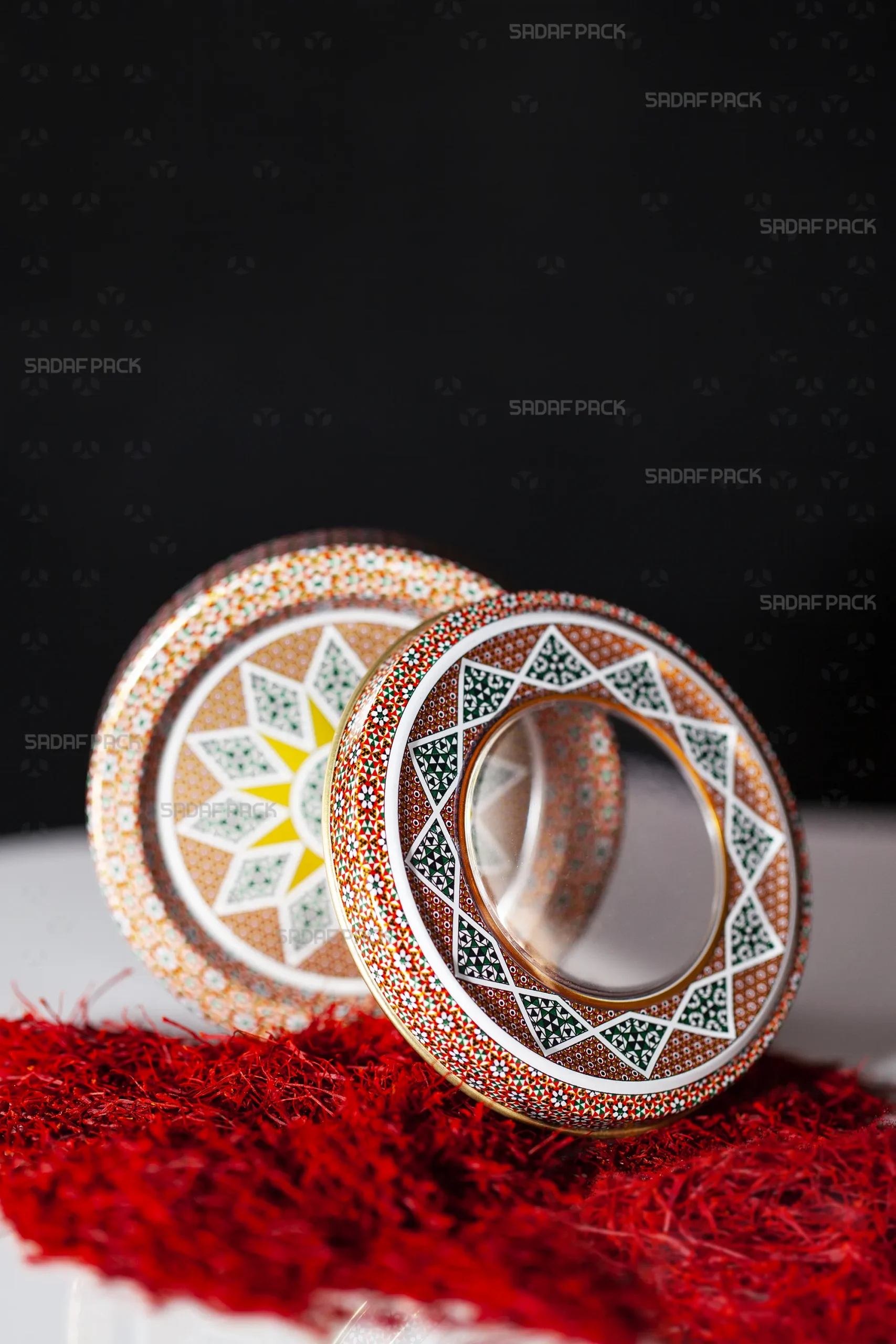 یک قوطی 5 گرم فلزی صدف پک با طرح خاتم قرمز محصول شرکت صدف پک، که درب پنجرهای شفاف آن در جلو و بدنه طرحدار آن در عقب، بر بستری از زعفران سرگل نمایش داده شدهاند.