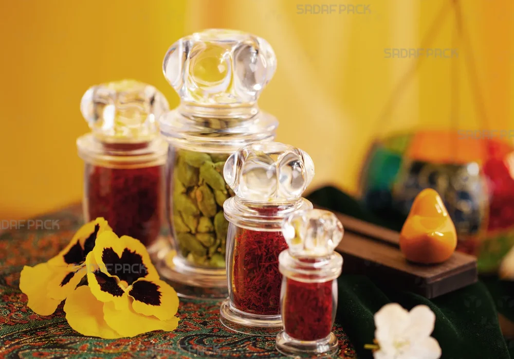 چیدمان هنری چهار سایز مختلف از ظروف کریستالی سری میلاد صدف پک، پر شده با زعفران و هل، در کنار گل‌های بنفشه زرد و پارچه سنتی ترمه که حس لوکس و اصیل ایرانی را القا می‌کند.