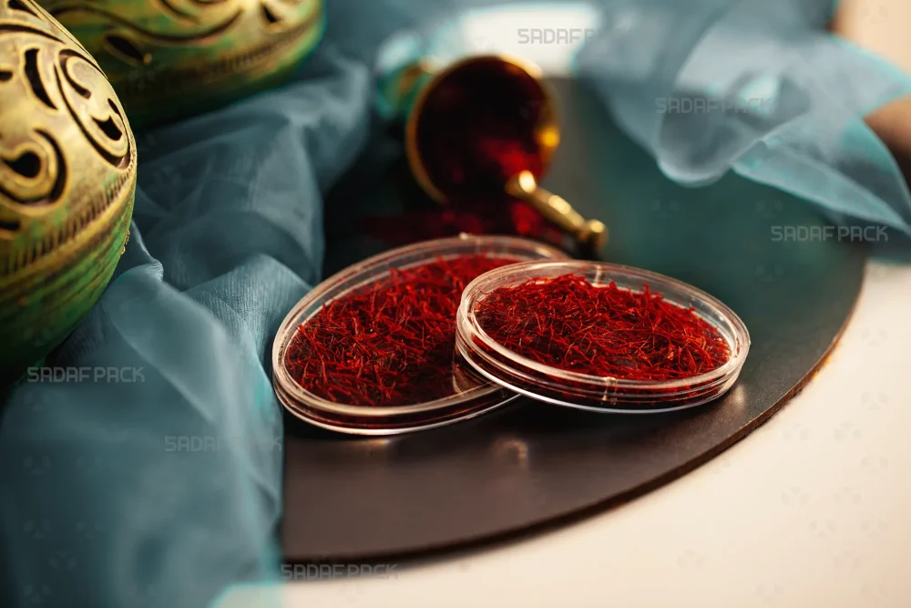 ظروف کریستالی بیضی ۳ و ۴ صدف پک، پر شده با زعفران صادراتی در یک چیدمان هنری با پارچه آبی فیروزه‌ای.