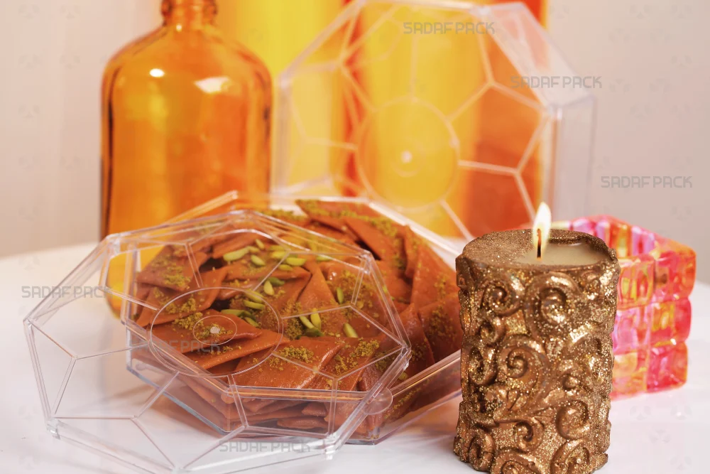 ظرف کریستالی هشت ضلعی صدف پک باز شده و پر از سوهان تازه با تزئین خلال پسته، در کنار یک شمع طلایی روشن برای نمایش کاربرد در بسته‌بندی شیرینی‌جات صادراتی